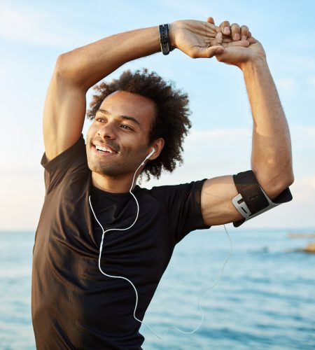 Portrait of a smiling afro-american sports man stretching his muscular arms before workout by the sea, using music app on his smartphone. Dark-skinned athlete warming up before running.